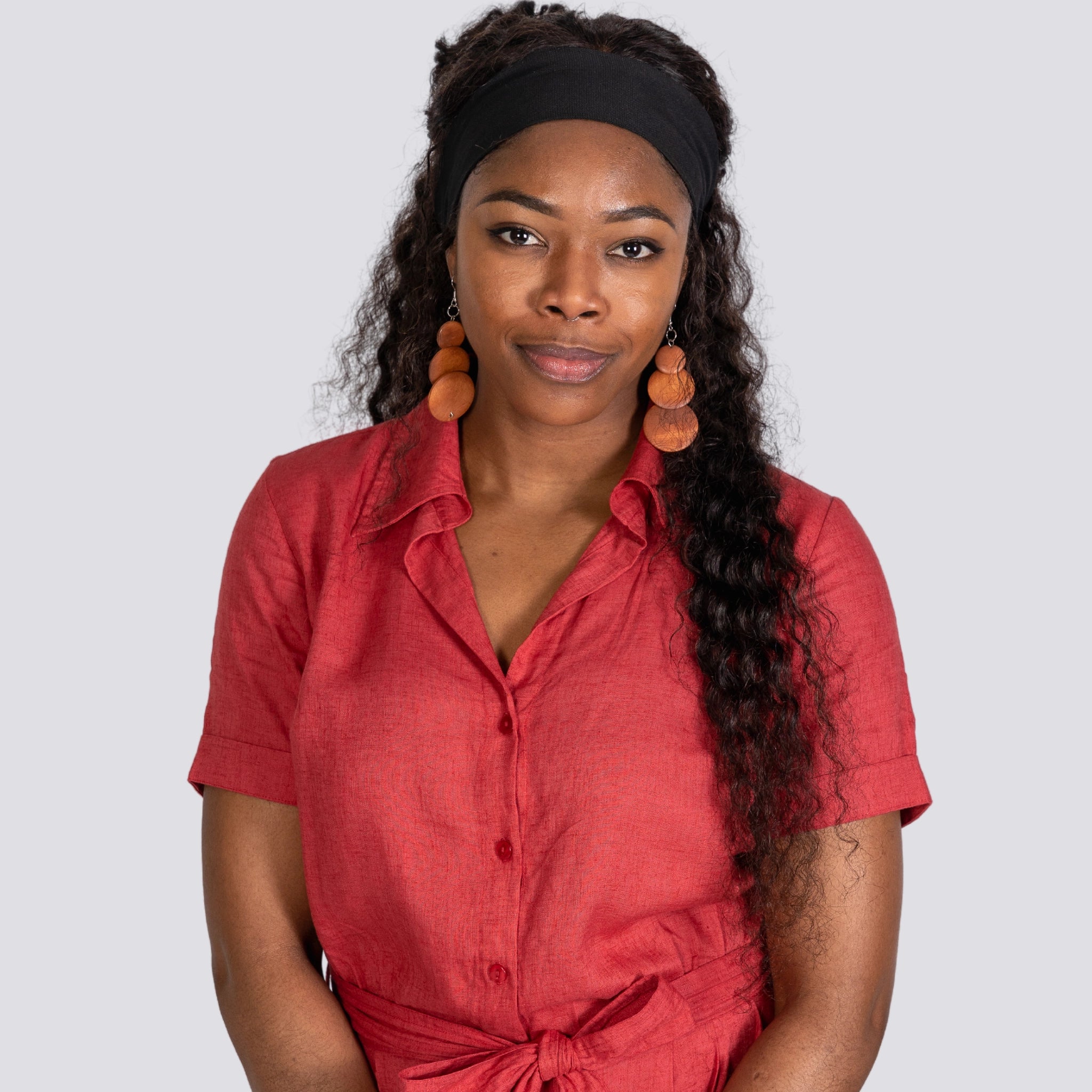 A woman in a Karee Milano Red Serenity Viscose Linen Jumpsuit and black headband, wearing round wooden earrings, looks directly at the camera with a neutral expression.