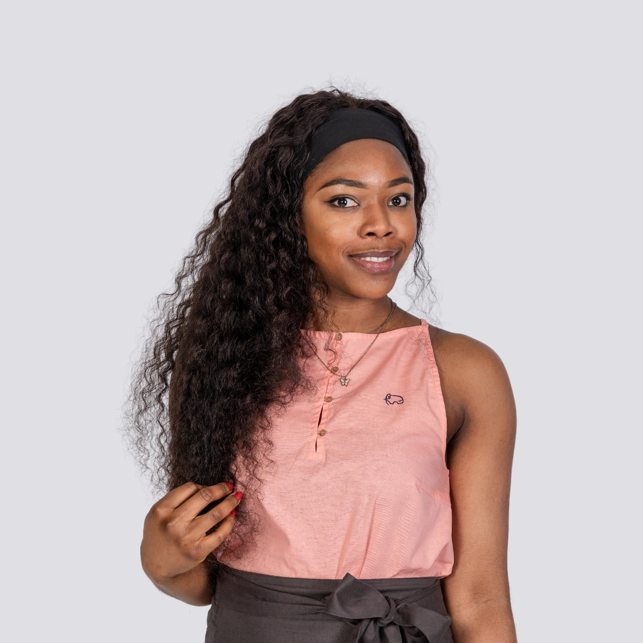A woman with long curly hair, wearing a Karee Wild Rose Harmony Halter Cami Top made of sustainable materials, and a headband, standing against a grey background, looking slightly to her left.