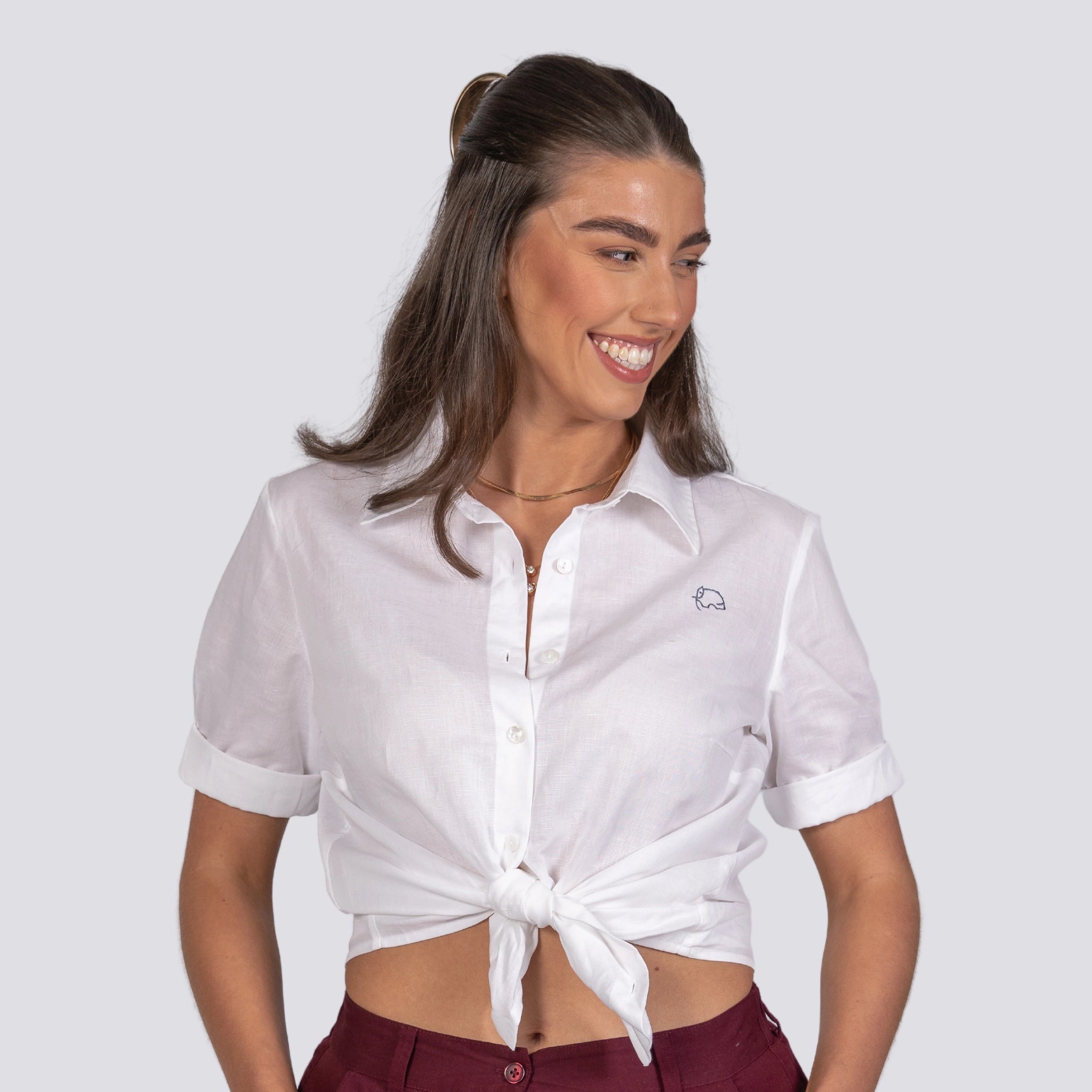 Woman smiling, wearing a knotted Karee Pure Elegance White Linen Cotton Shirt and maroon pants, with a minimalist necklace, against a gray background.