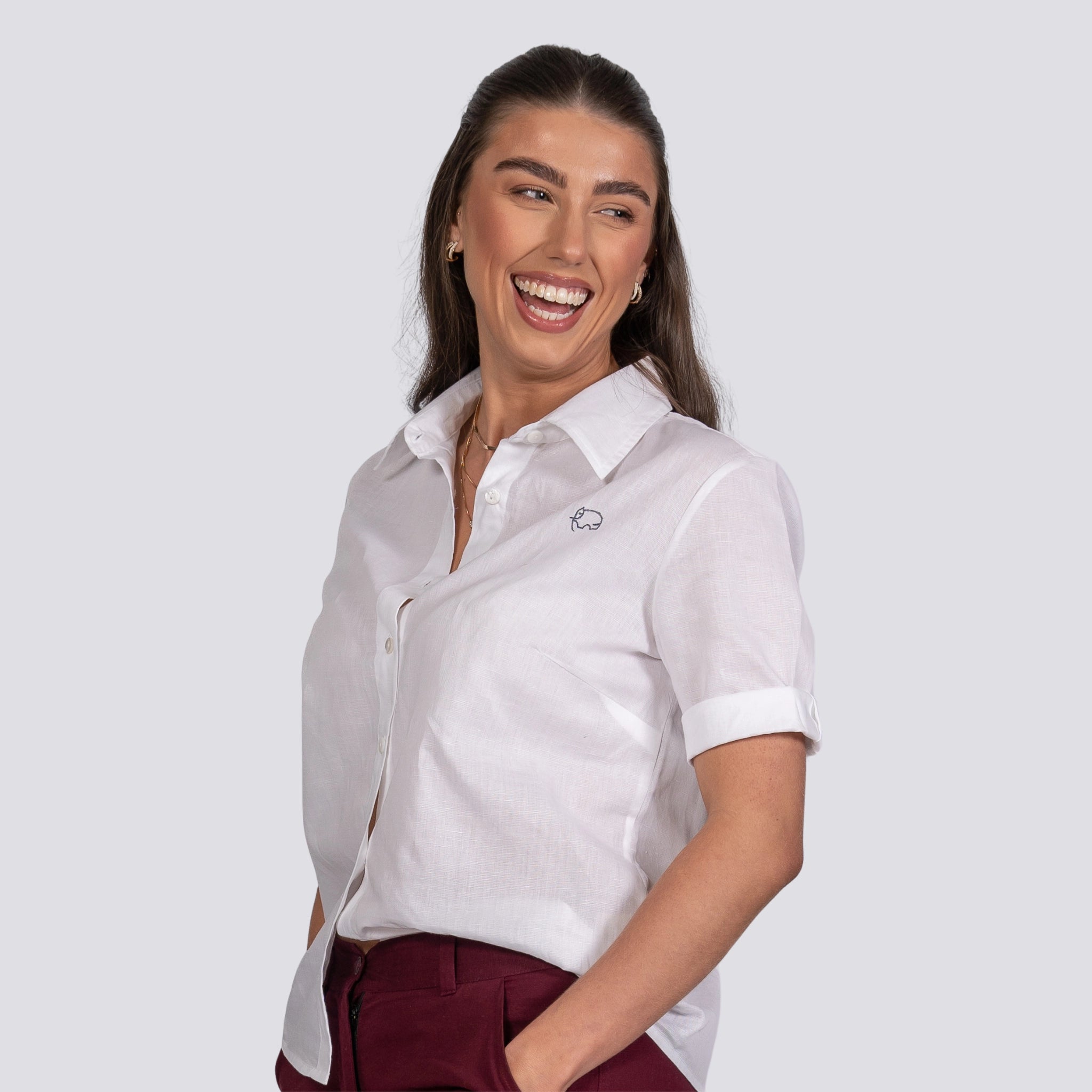 A woman with long dark hair, wearing a white Karee Pure Elegance White Linen Cotton Shirt and burgundy pants, smiling joyfully at something off-camera.