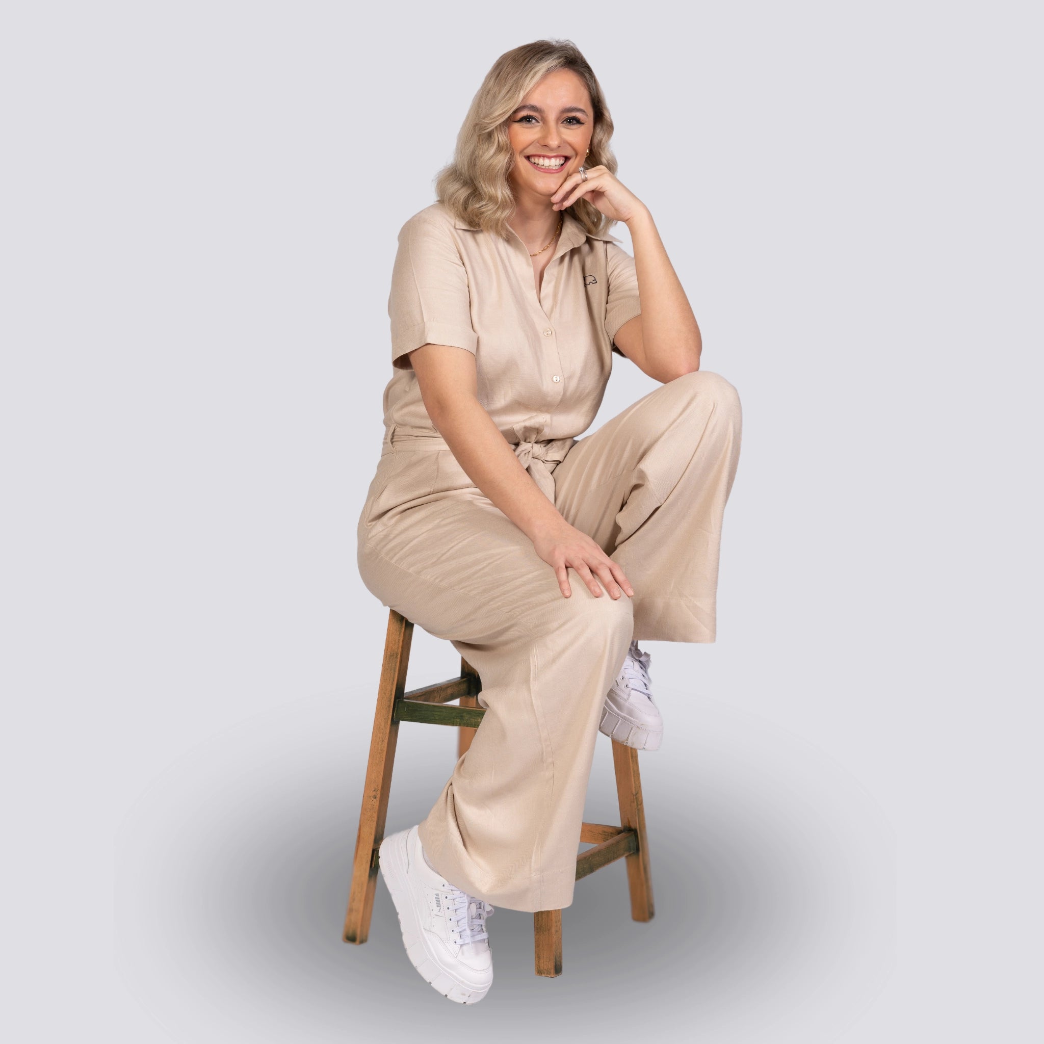 Smiling woman in a Karee Merino Elegance Viscose Linen Jumpsuit sitting on a wooden stool against a gray background.