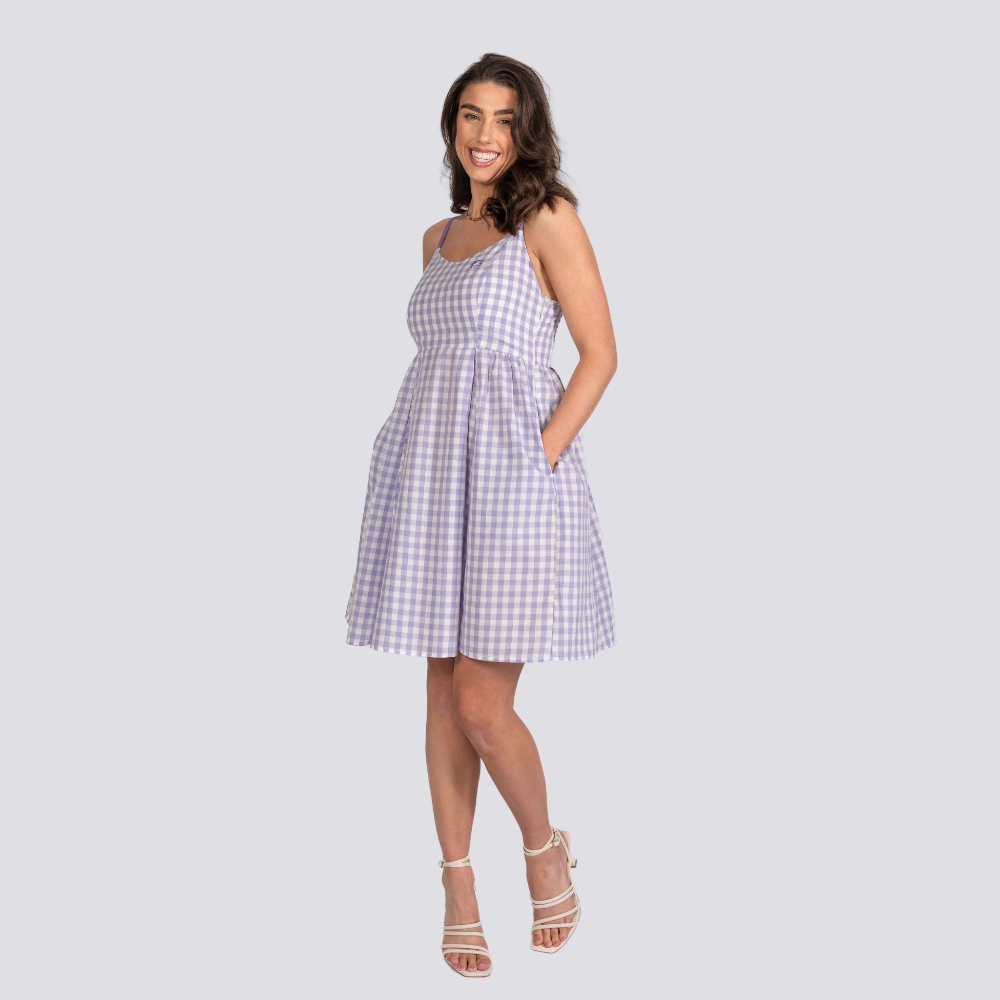 Woman in a Karee lavender plaid cotton mini dress and white sandals, smiling and posing against a grey background.