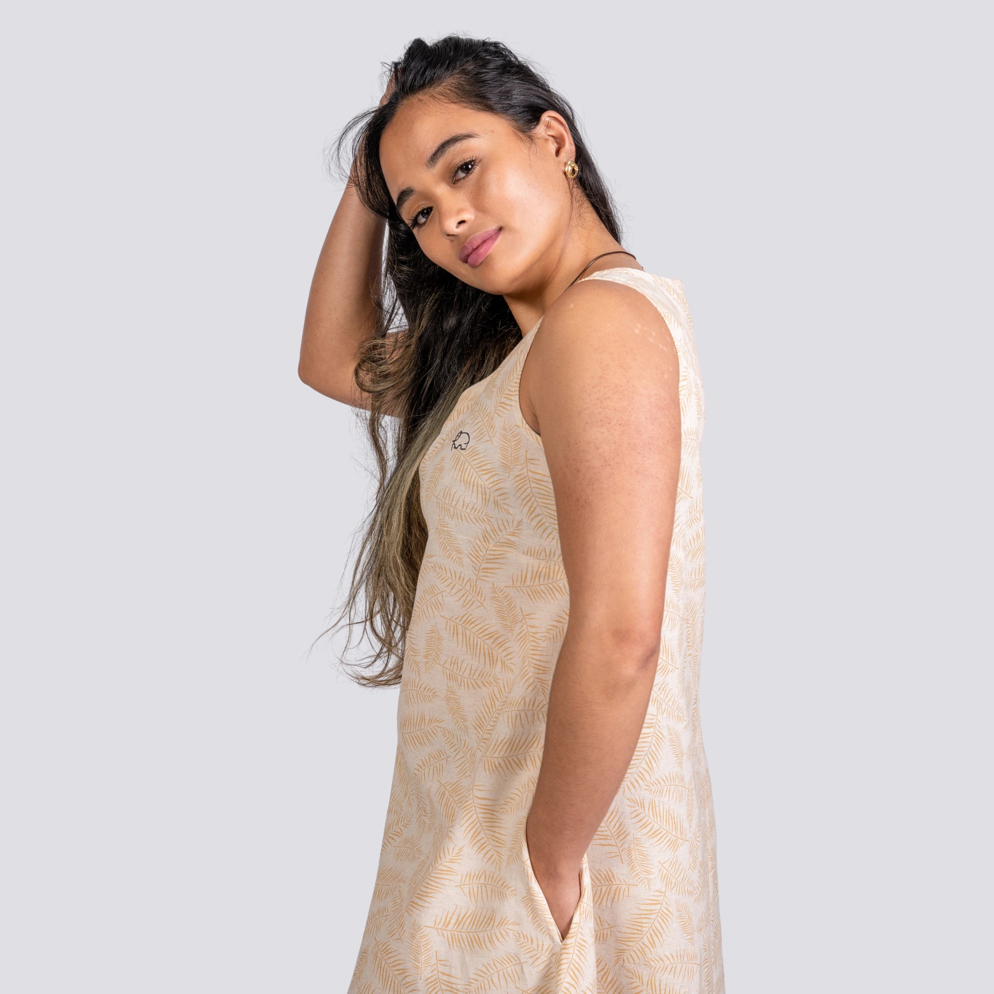 A woman in a beige, sleeveless U-neck Karee Mystic Serenity High Low Linen Cotton Midi Dress with a feather pattern looks over her shoulder, with a hand in her hair, against a light gray background.