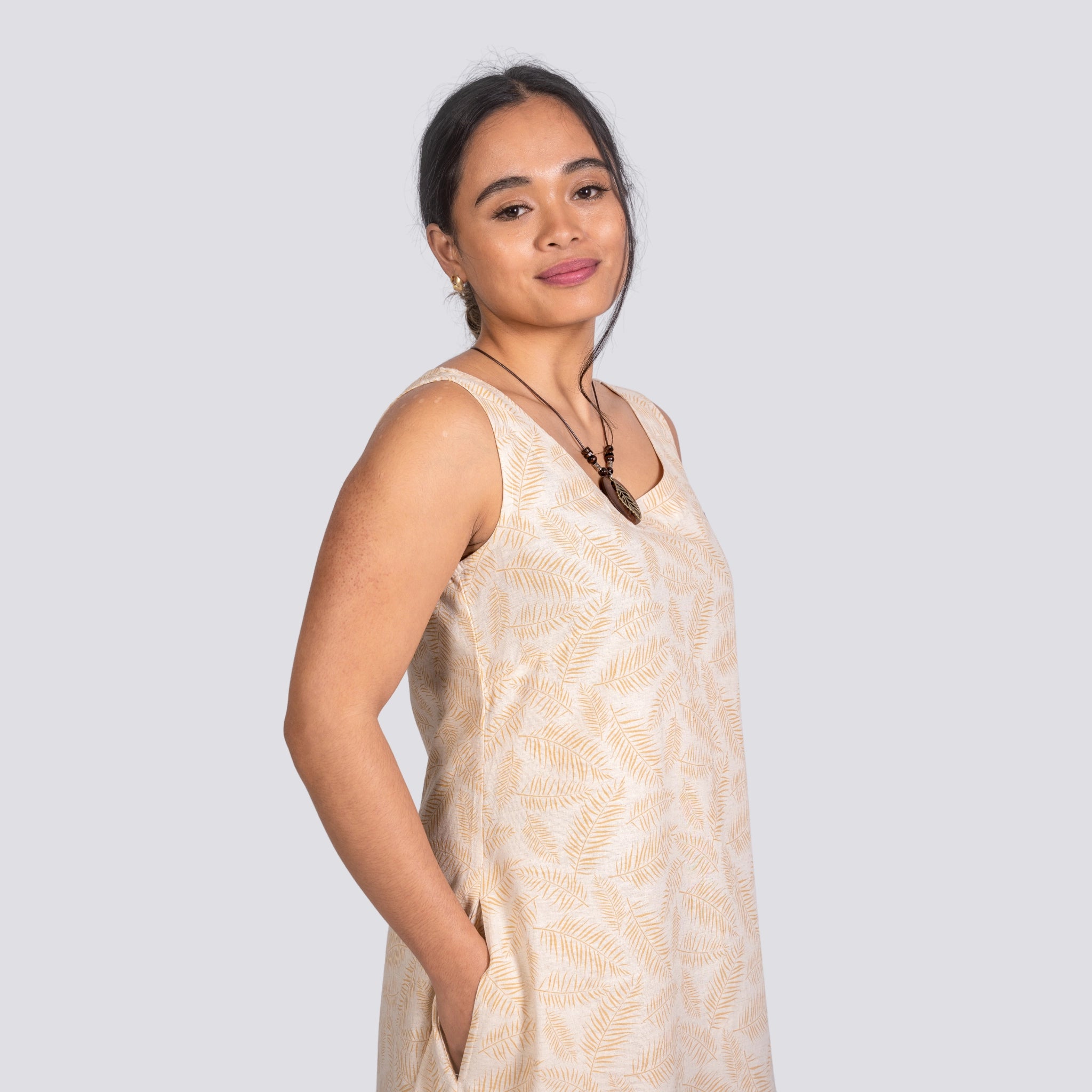 A young woman wearing a Karee Mystic Serenity High Low Linen Cotton Midi Dress with a palm leaf pattern, standing sideways, smiling subtly at the camera.