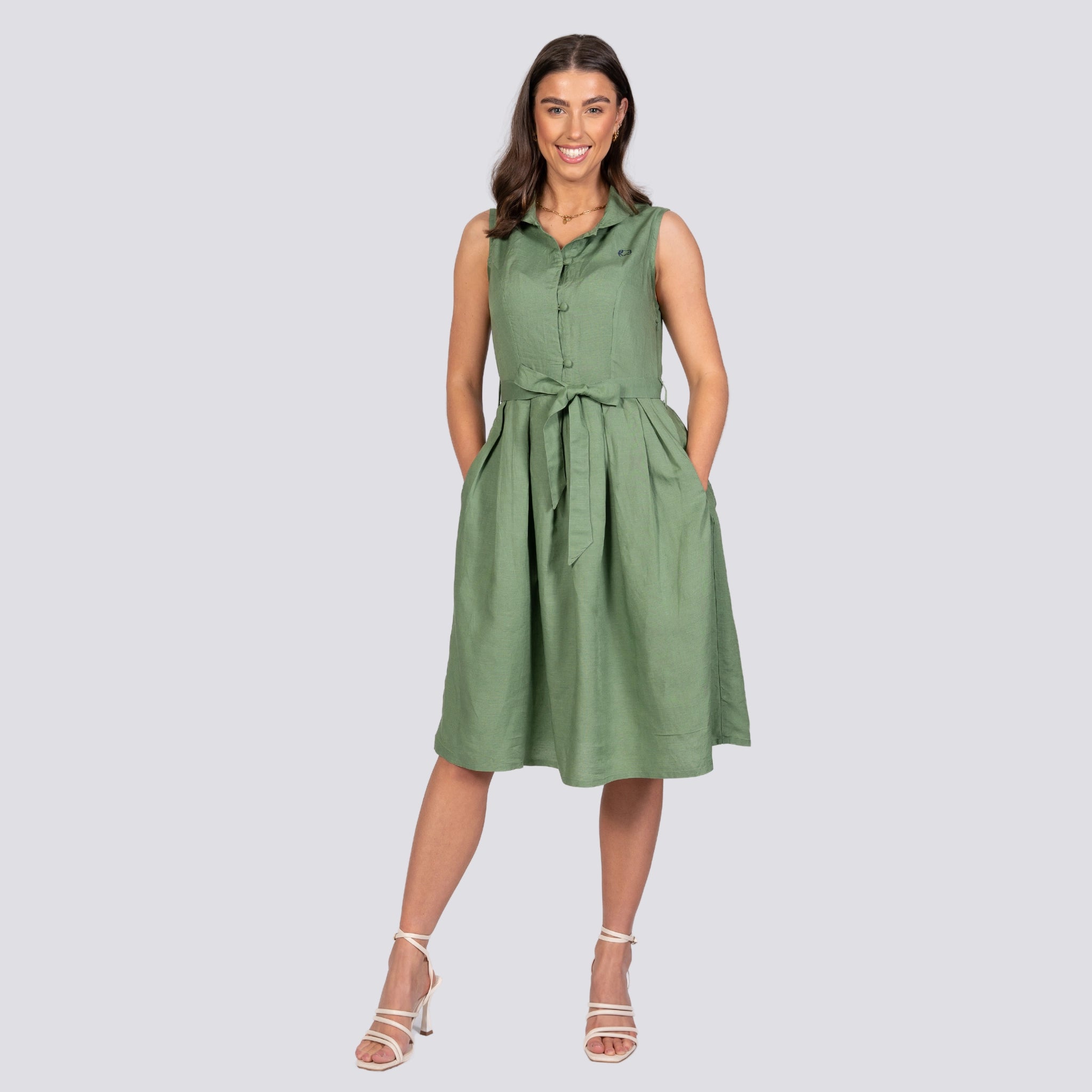 Woman in a green sleeveless Karee Vintage Midi Button-Up Dress For Women with a tie waist, smiling and standing against a plain gray background. She wears white heeled sandals.