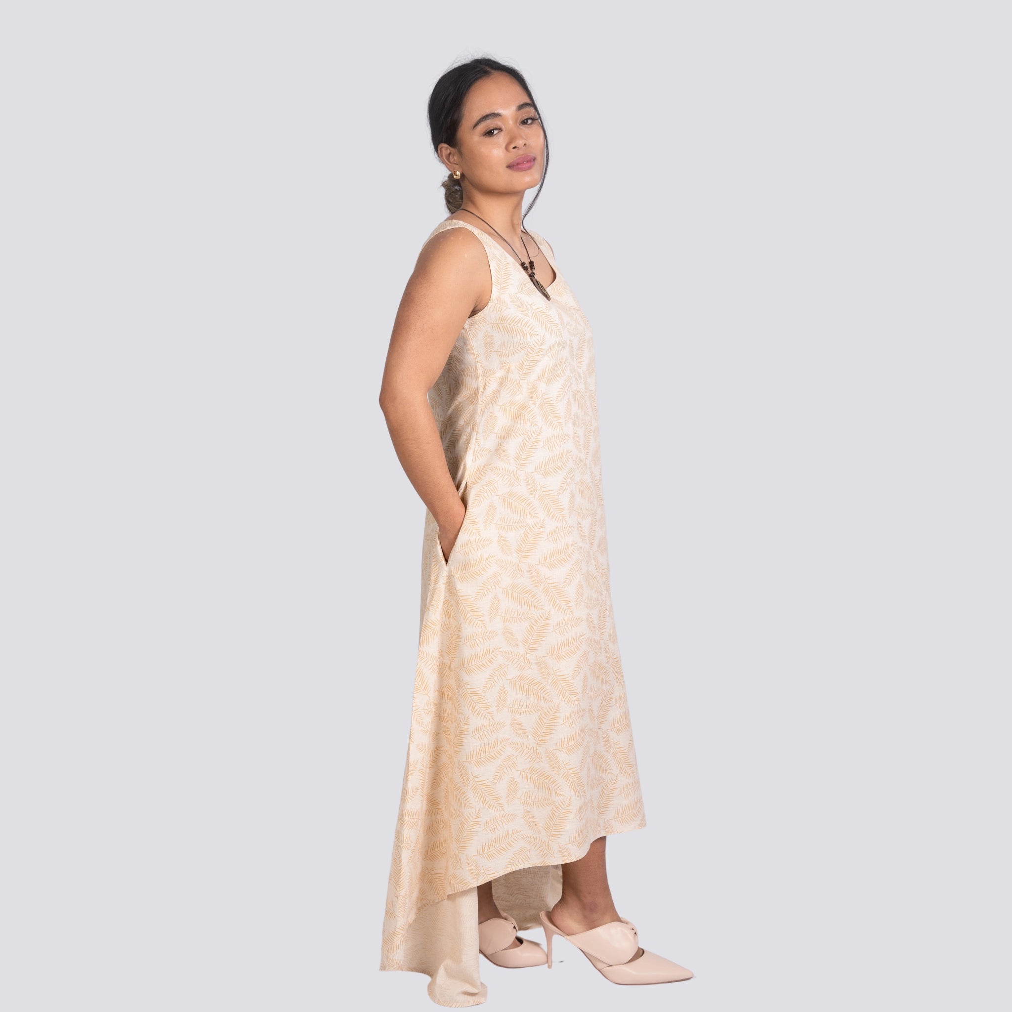 Woman in an elegant beige Karee High Low Linen Cotton Midi Dress and white shoes, standing sideways and looking over her shoulder, against a light gray background.