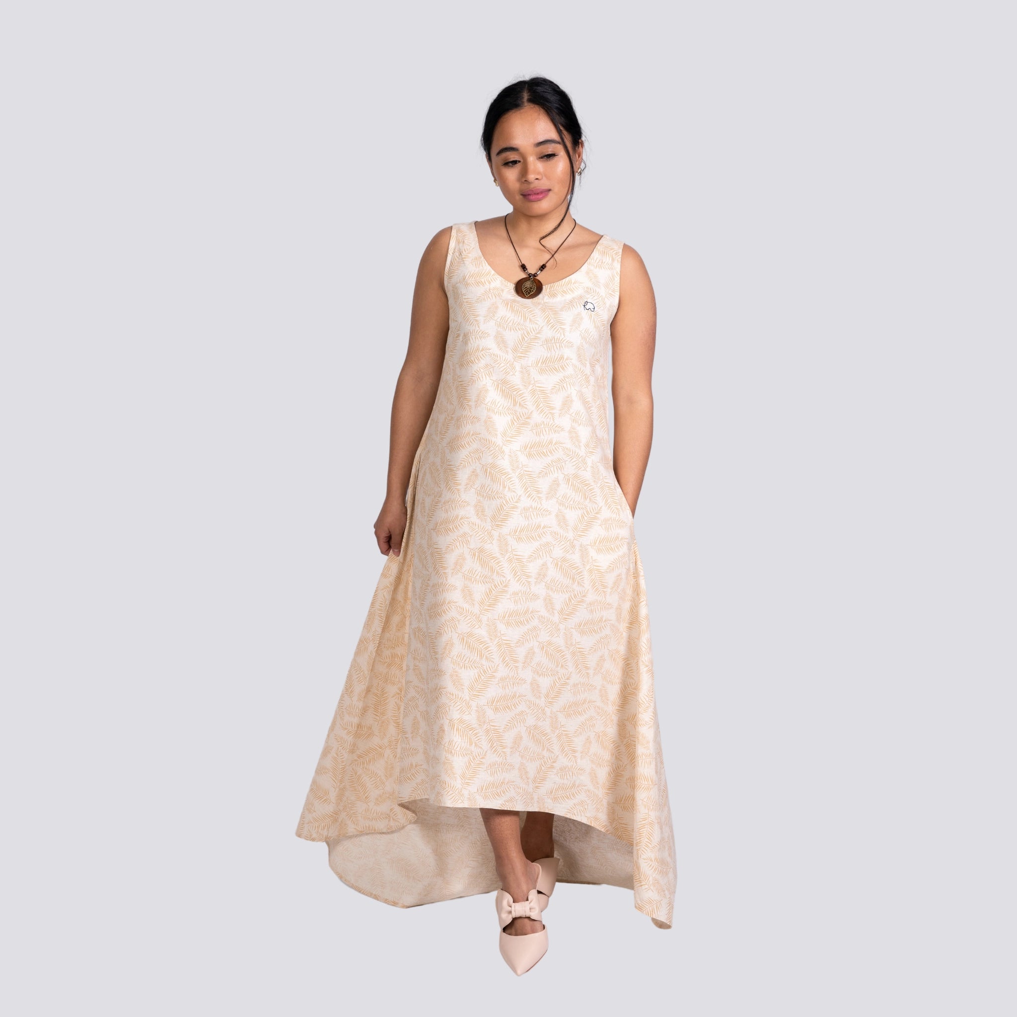 A woman in a flowing beige Karee Mystic Serenity High Low Linen Cotton Midi Dress and light pink shoes stands with closed eyes, against a grey background.