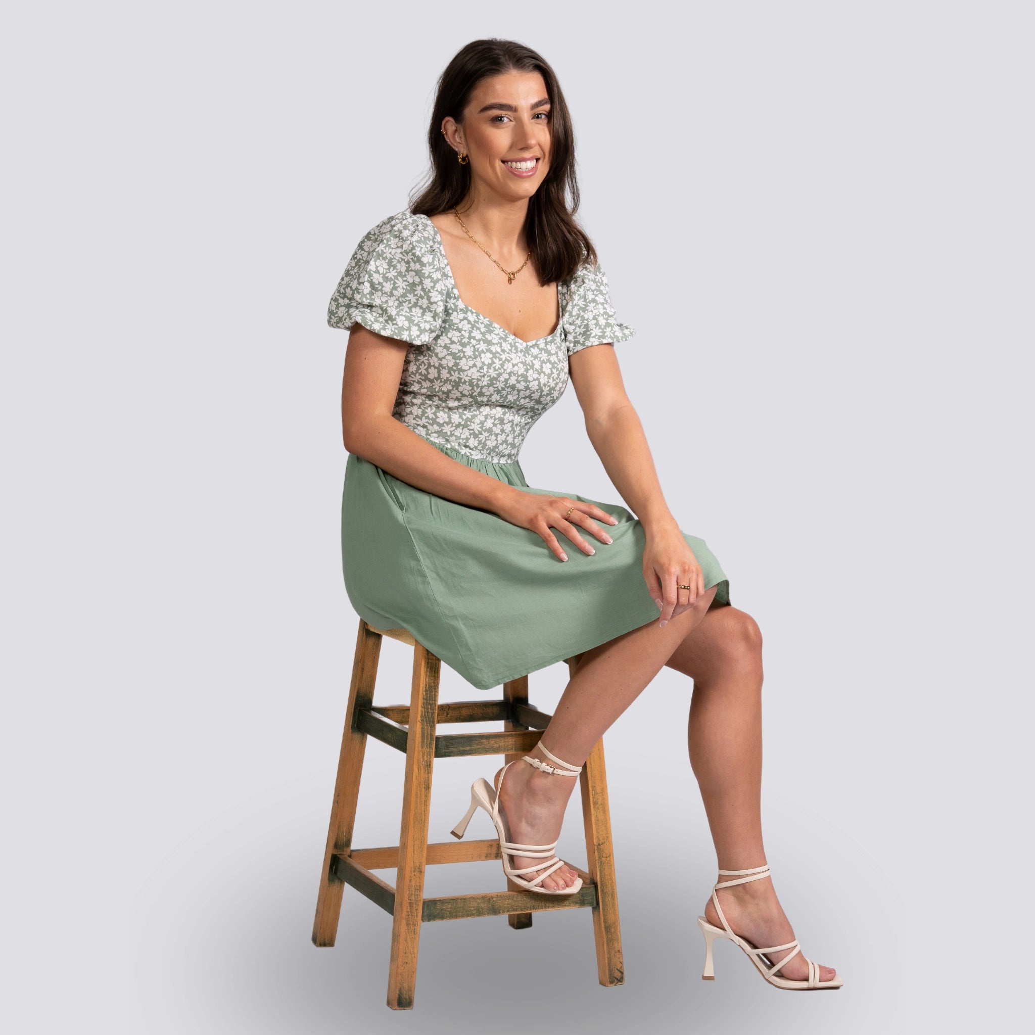 Woman in a Sirocco Blooms Green Frock Dress by Karee with a knee-length hemline and floral top sitting on a wooden stool with a light gray background.
