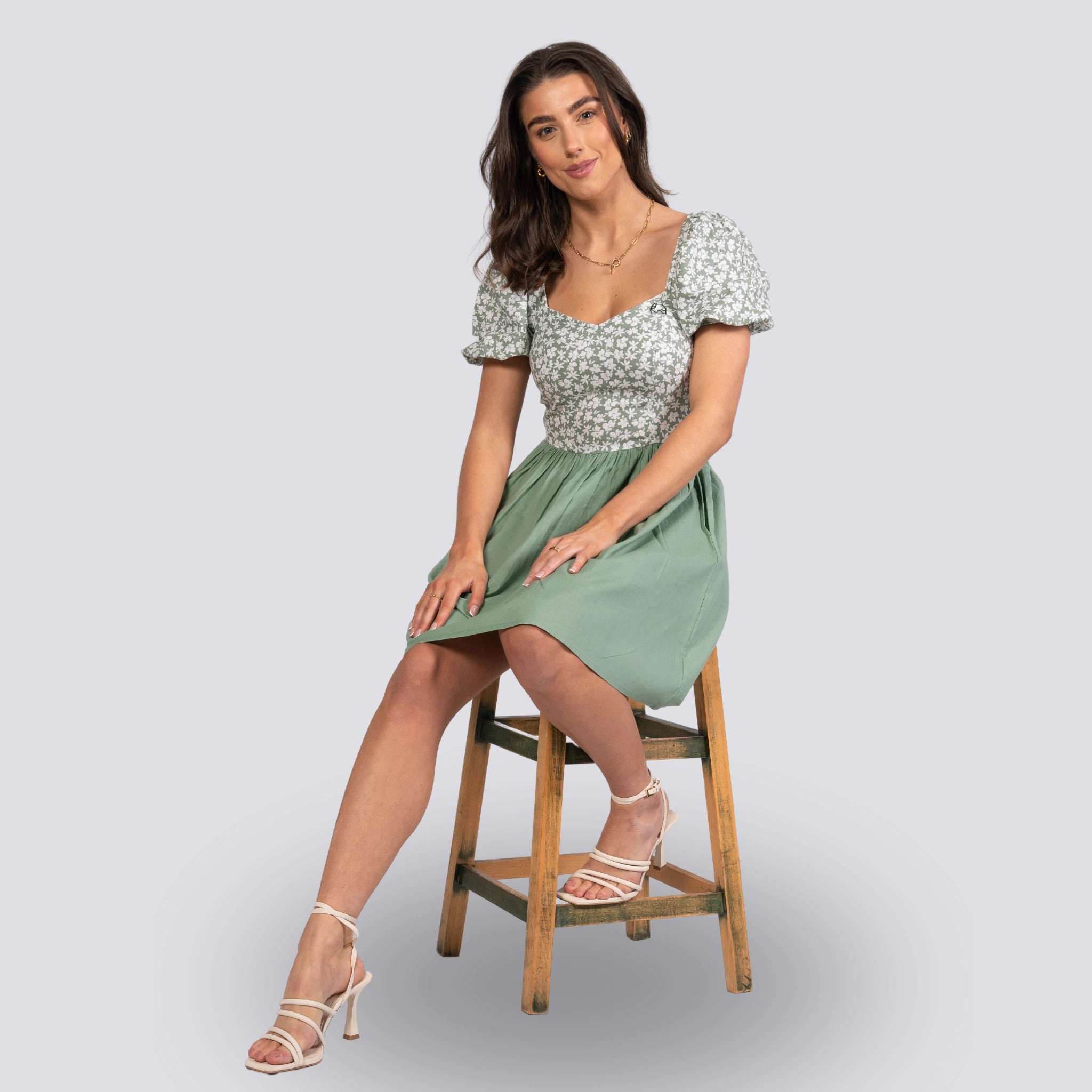 A woman in a Karee Sirocco Blooms green frock dress with a knee-length hemline sits on a wooden stool against a grey background, looking at the camera.