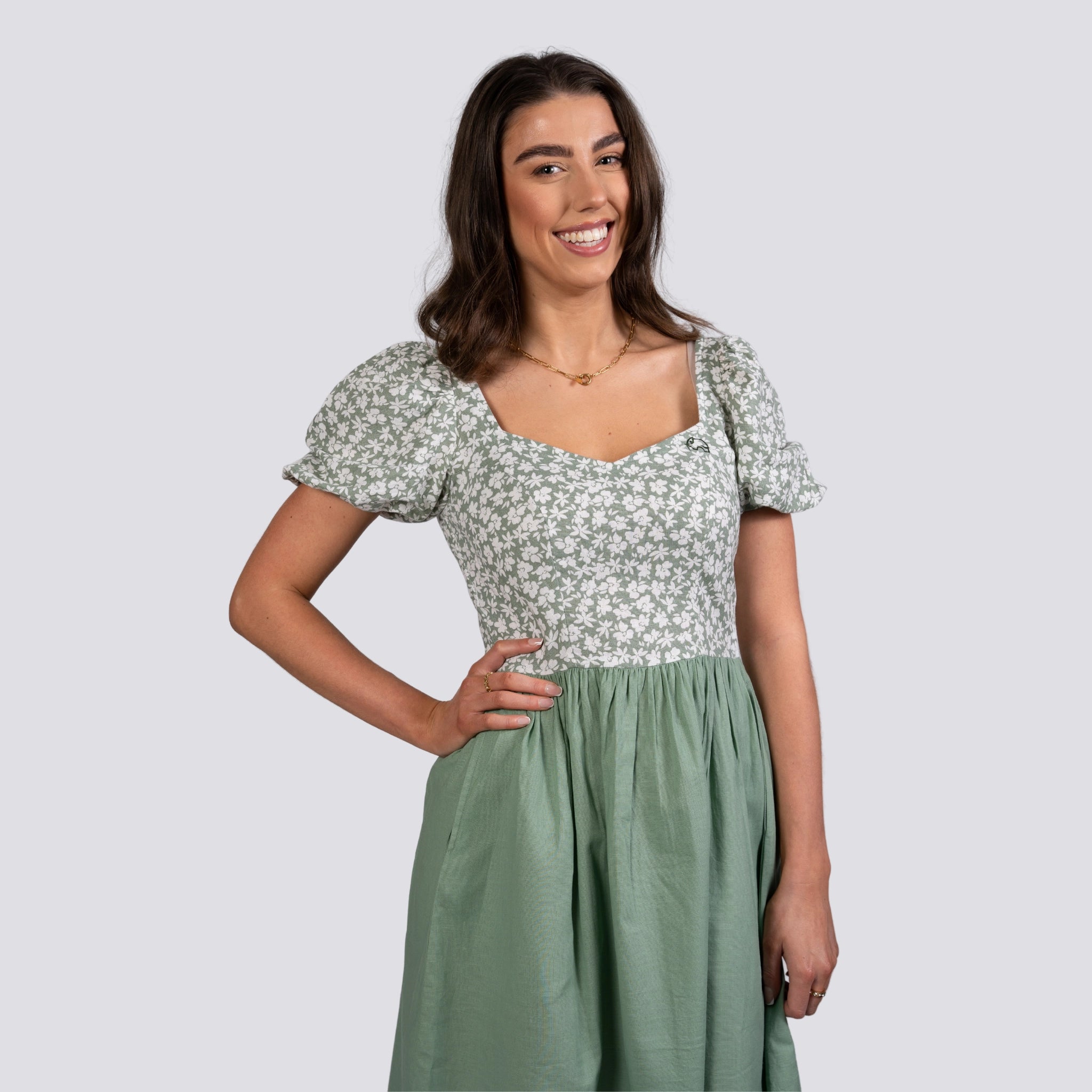 Young woman smiling, wearing a green and white Karee Frock Dress with a knee-length hemline, standing against a light gray background.