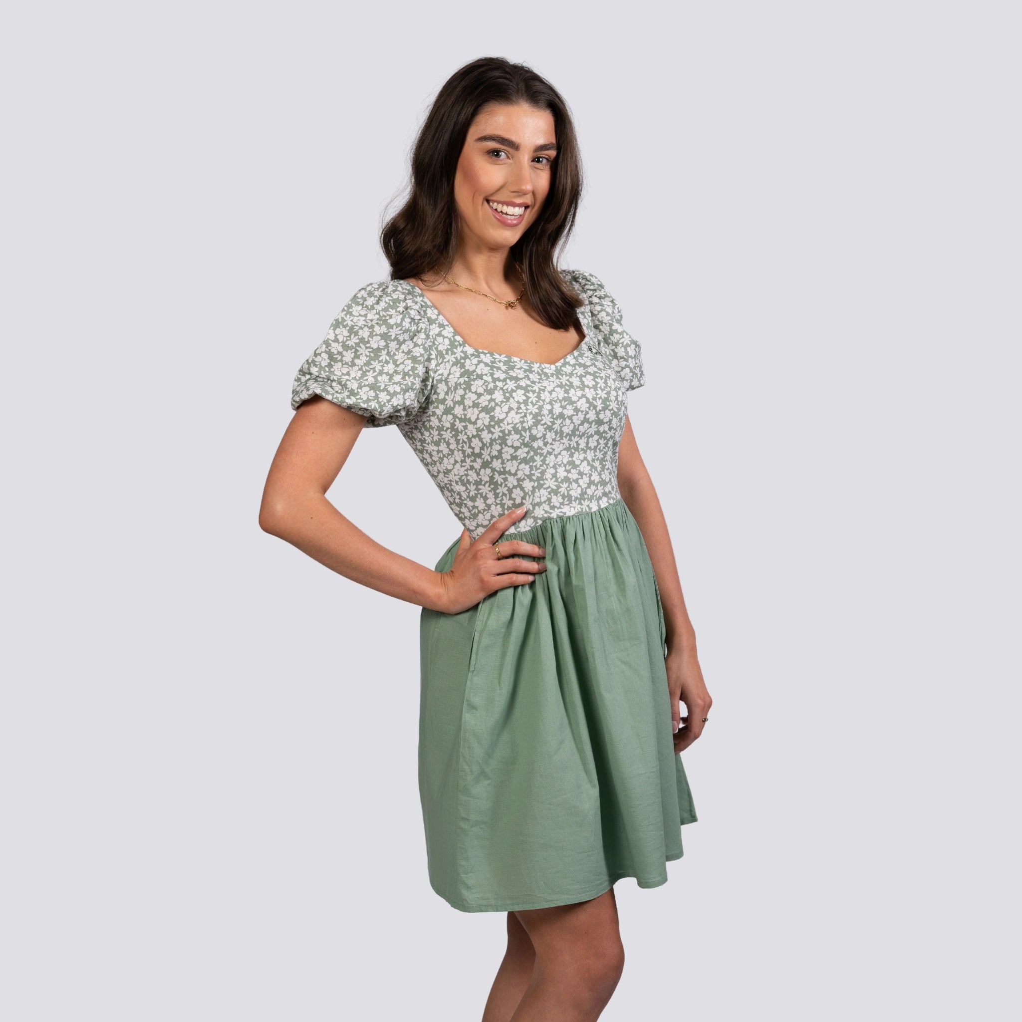 A woman in a Karee Sirocco Blooms Green Frock Dress smiles while posing with one hand on her hip against a gray background.