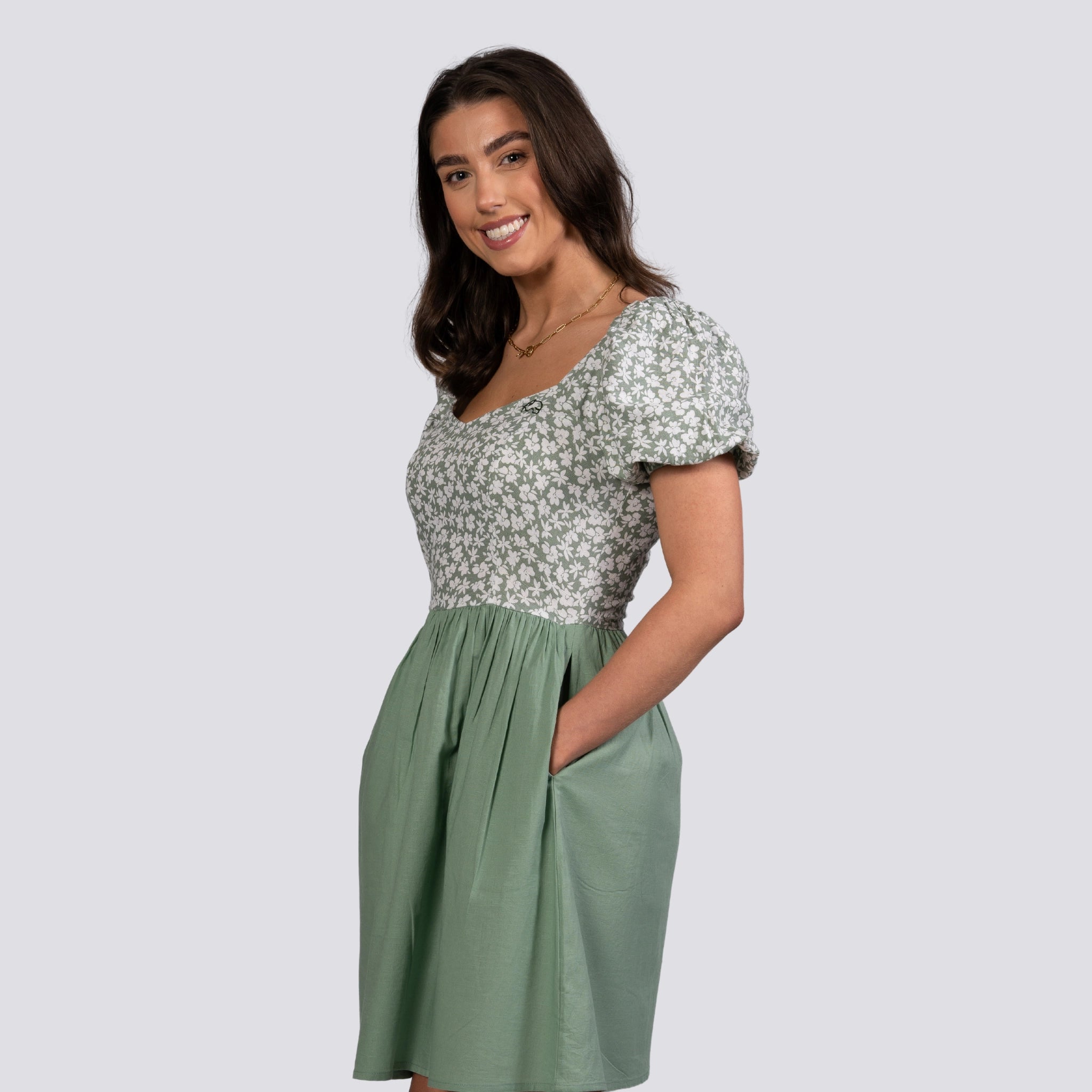A smiling young woman wearing a Karee Sirocco Blooms Green Frock Dress with a knee-length hemline, standing against a light gray background.
