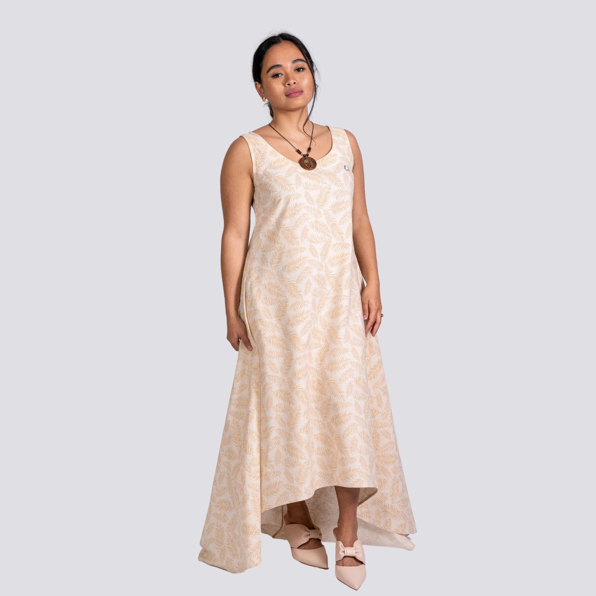 A woman in a sleeveless U-neck design beige Karee High Low Linen Cotton Midi Dress and beige shoes, standing against a gray background, looking at the camera.