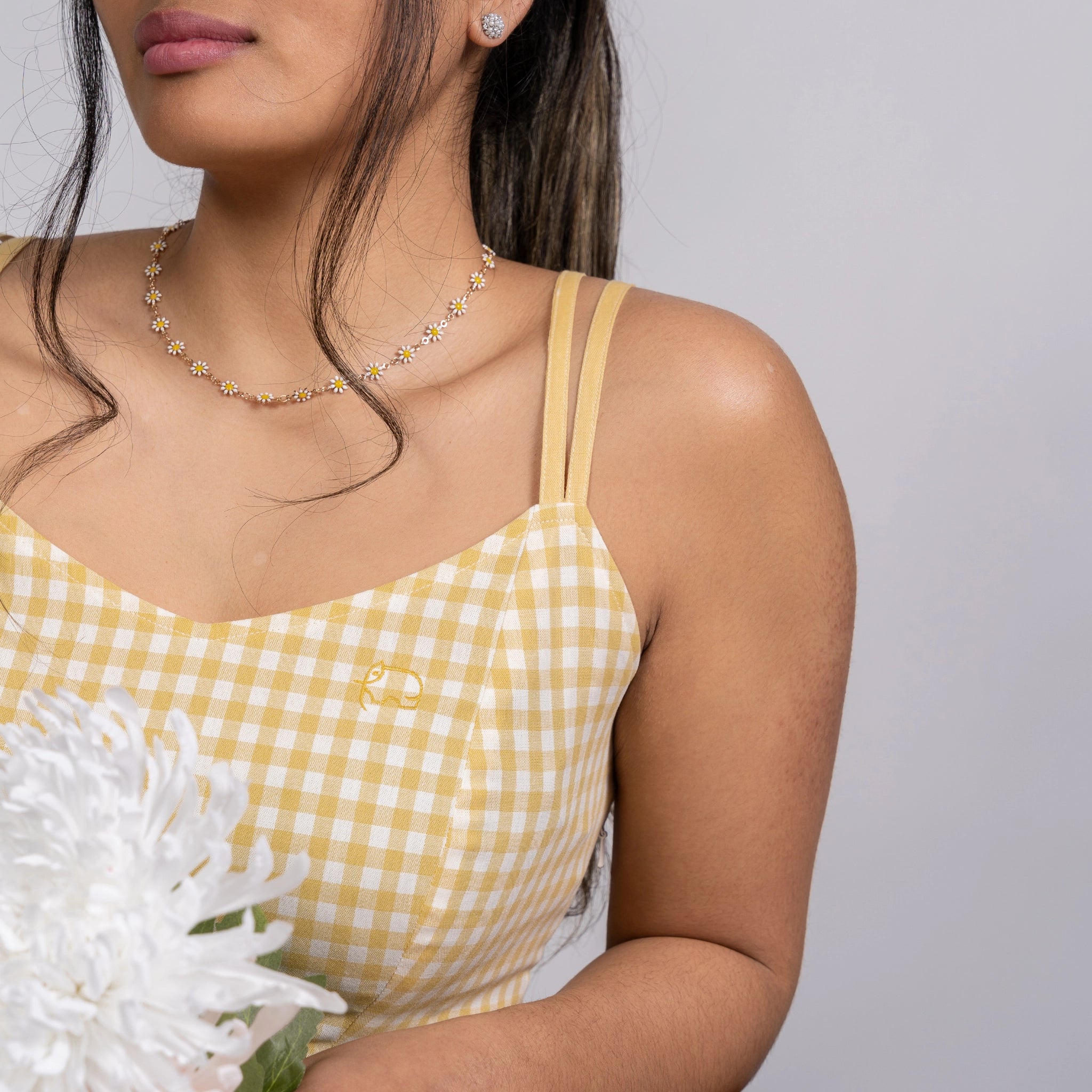 Woman wearing a Sunshine Chic Yellow Plaid Cotton Mini Dress by Karee and a floral necklace, holding a large white flower, cropped at shoulders.