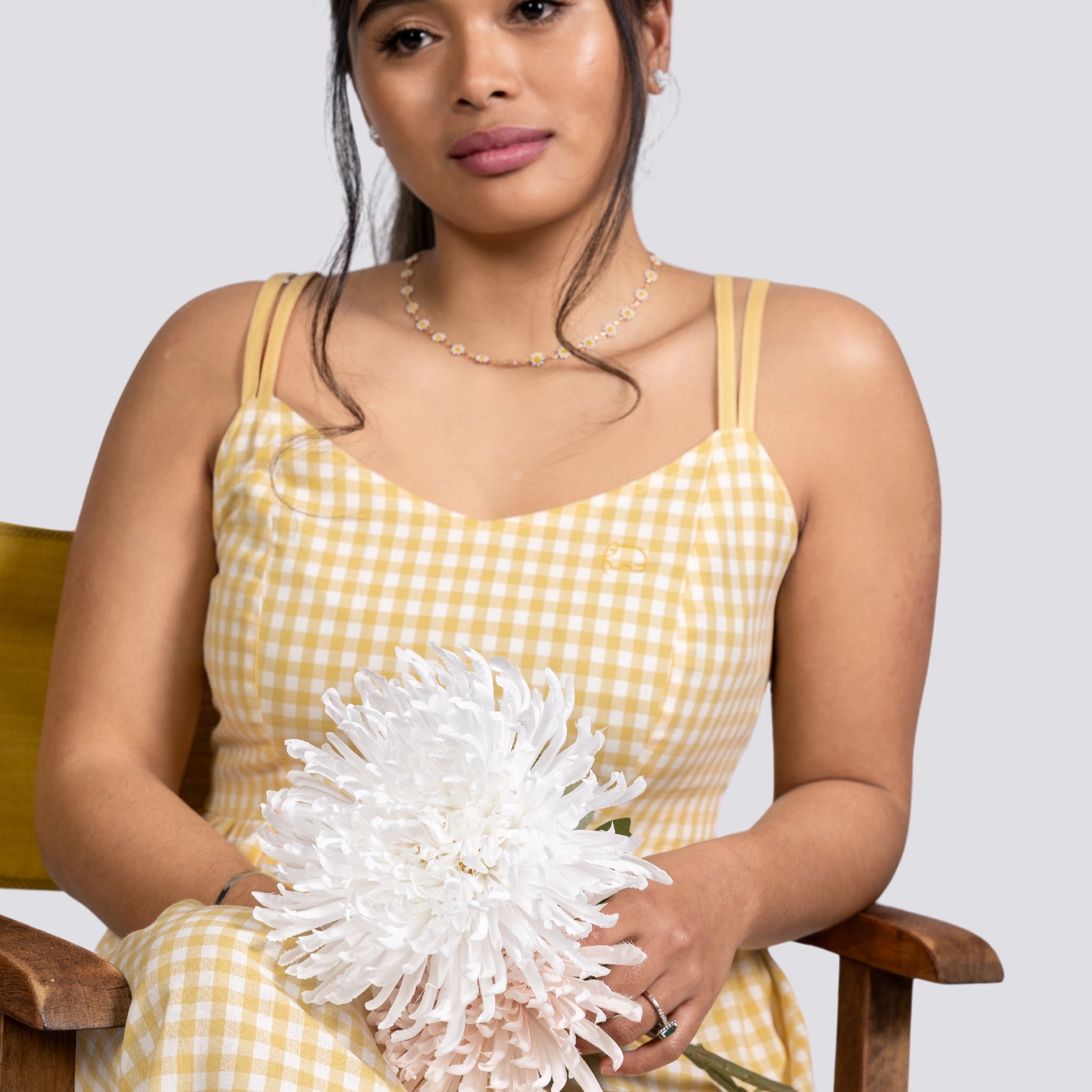 A woman in a Karee Sunshine Chic Yellow Plaid Cotton Mini Dress sitting on a chair, holding a large white chrysanthemum, looking at the camera.