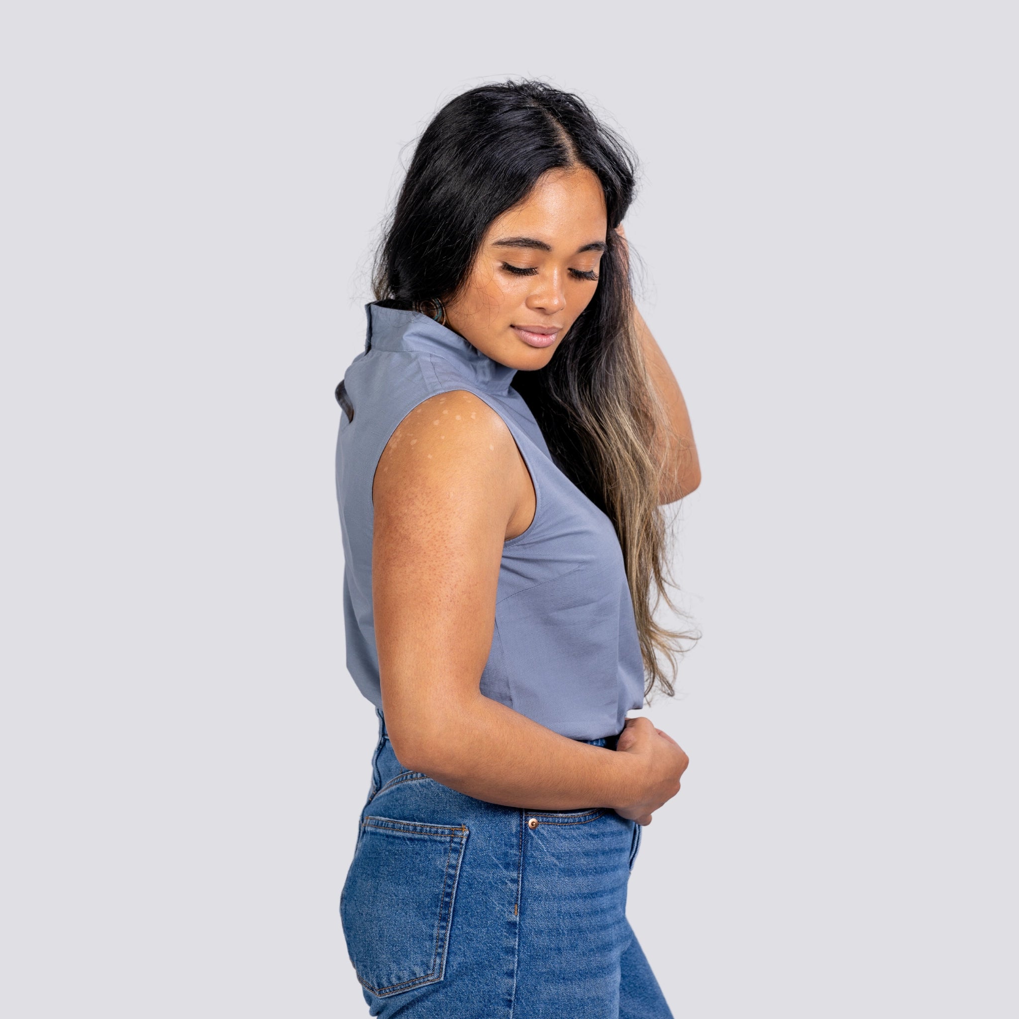 A woman in a Karee Linen Sleeveless Top in Classic Grey and jeans stands with her side to the camera, looking down and smiling slightly.