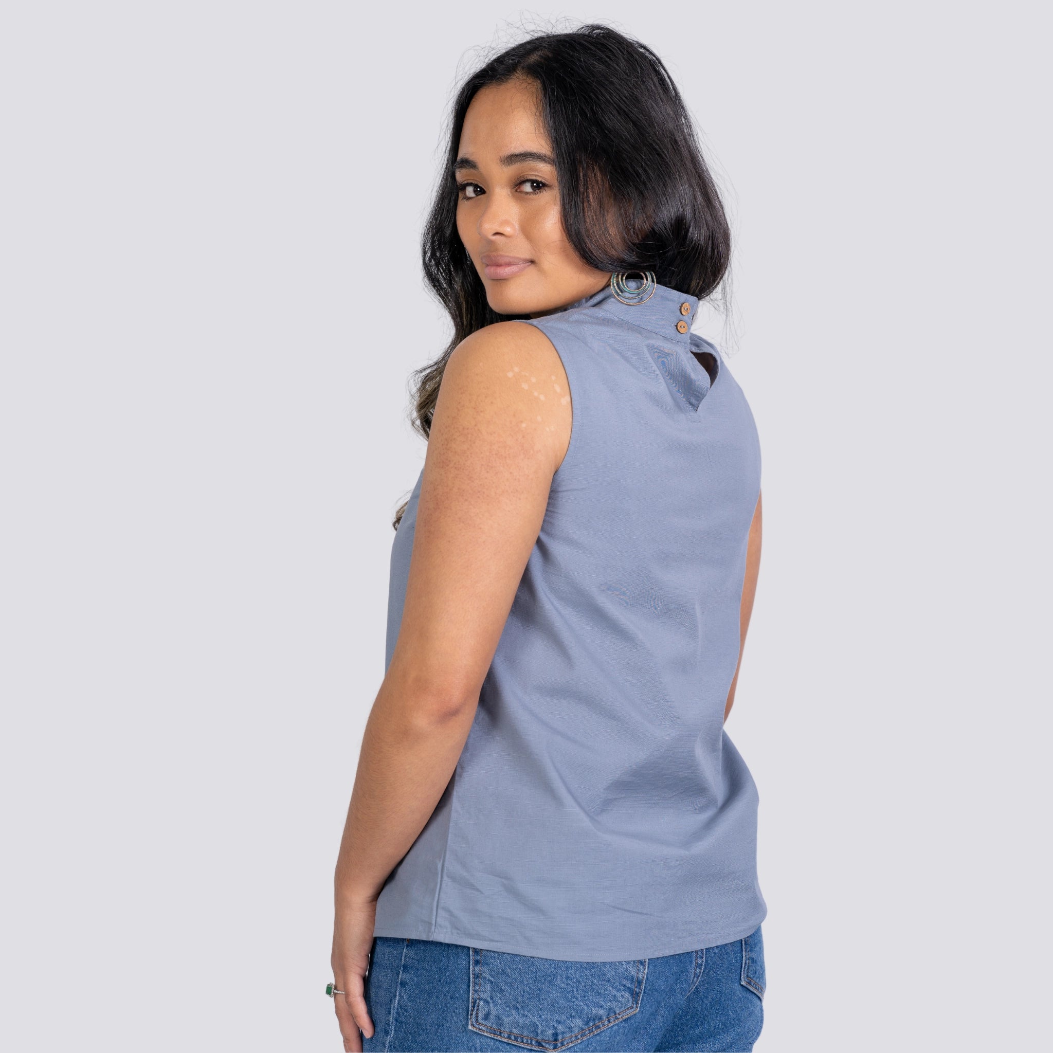 A woman in a Karee Linen Sleeveless Top in Classic Grey and jeans, looking over her shoulder with a subtle smile, against a plain light background.