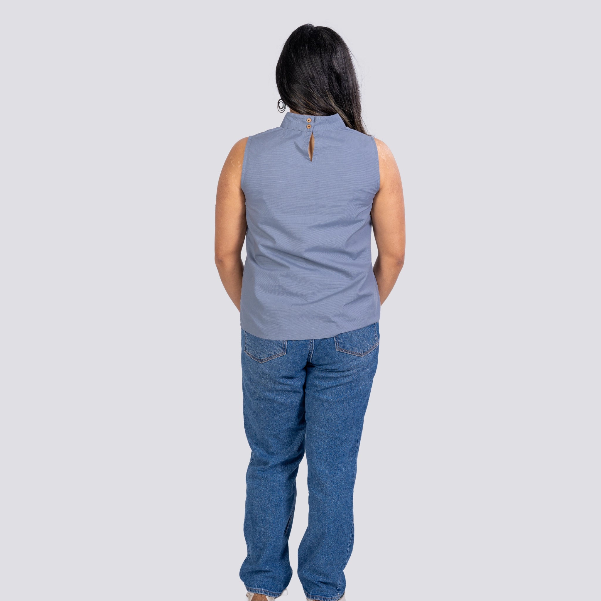 Woman standing with her back to the camera, wearing a sleeveless Karee Linen Top in Classic Grey and blue jeans, against a gray background.