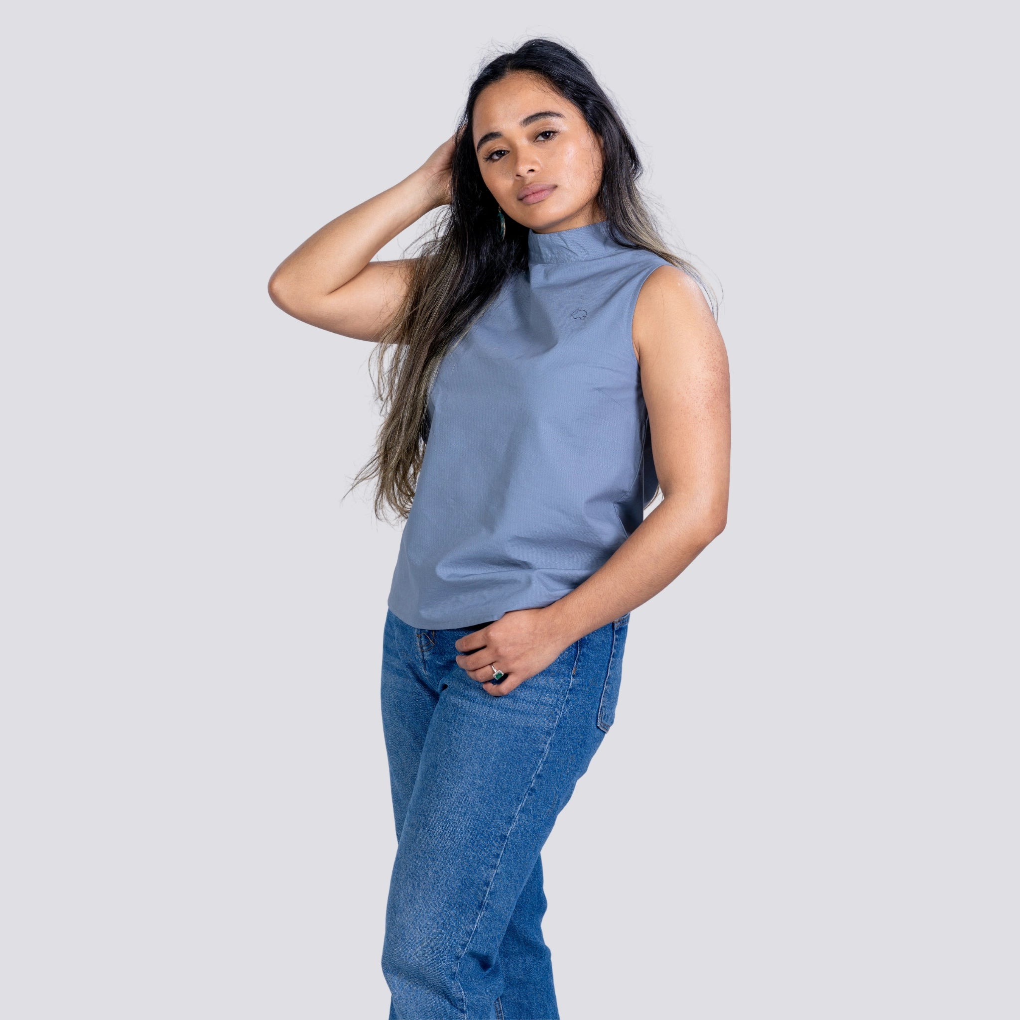 Woman in a sustainable fashion Karee Linen Sleeveless Top in Classic Grey and blue jeans posing with her hand in her hair, against a light gray background.