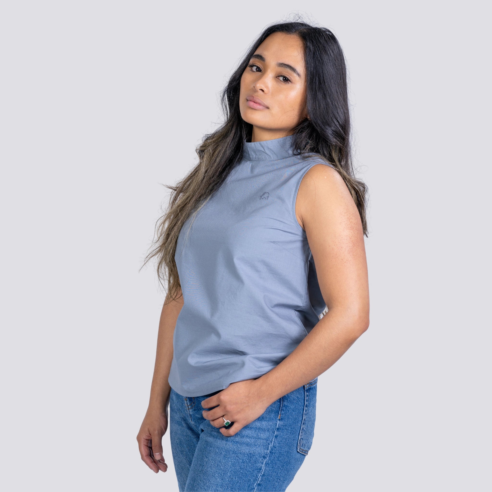 Woman in a Karee Linen Sleeveless Top in Classic Grey and blue jeans, standing with her hand on her hip, looking to the side against a light gray background.