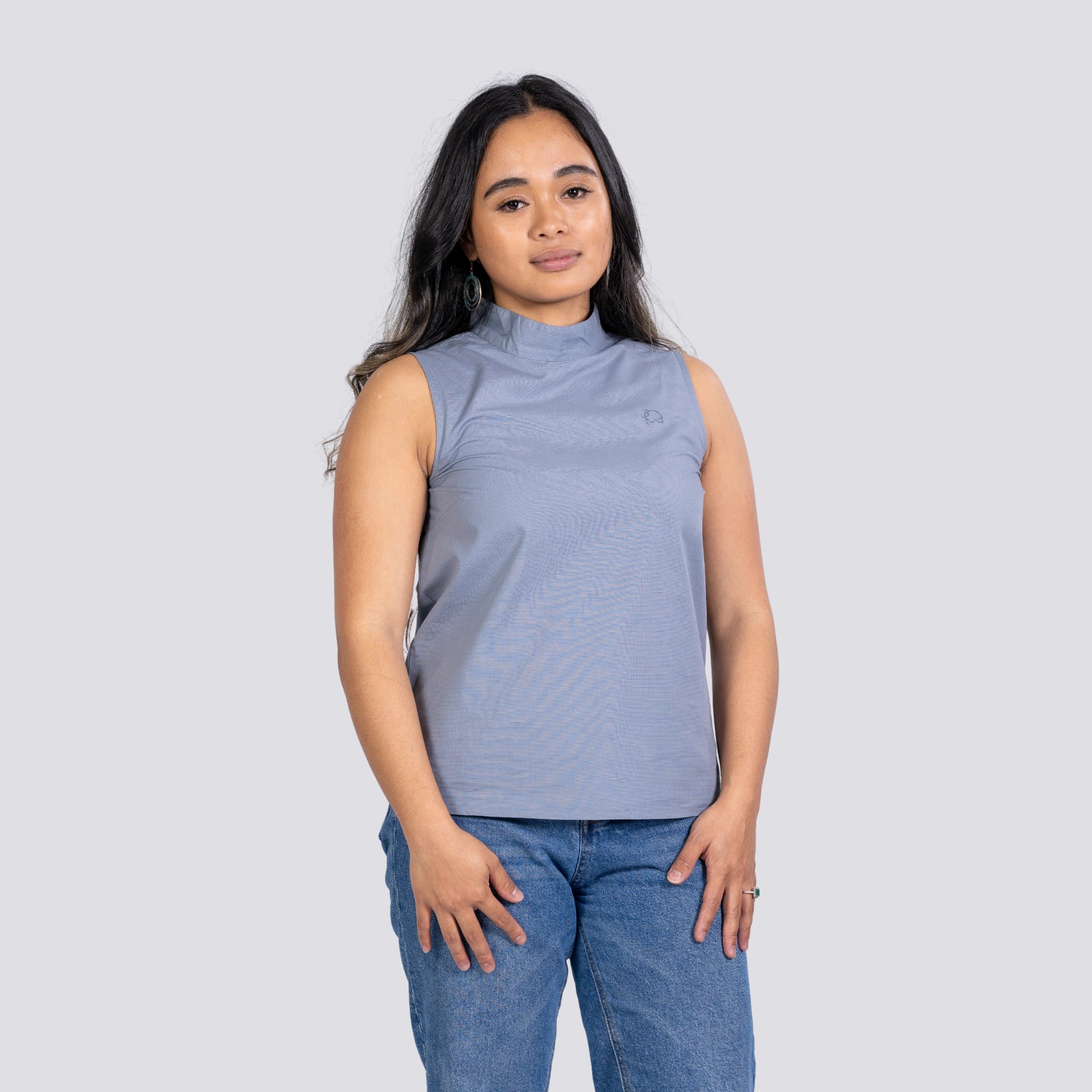 A young woman in a sustainable fashion Karee Linen Sleeveless Top in Classic Grey and blue jeans standing against a light gray background.