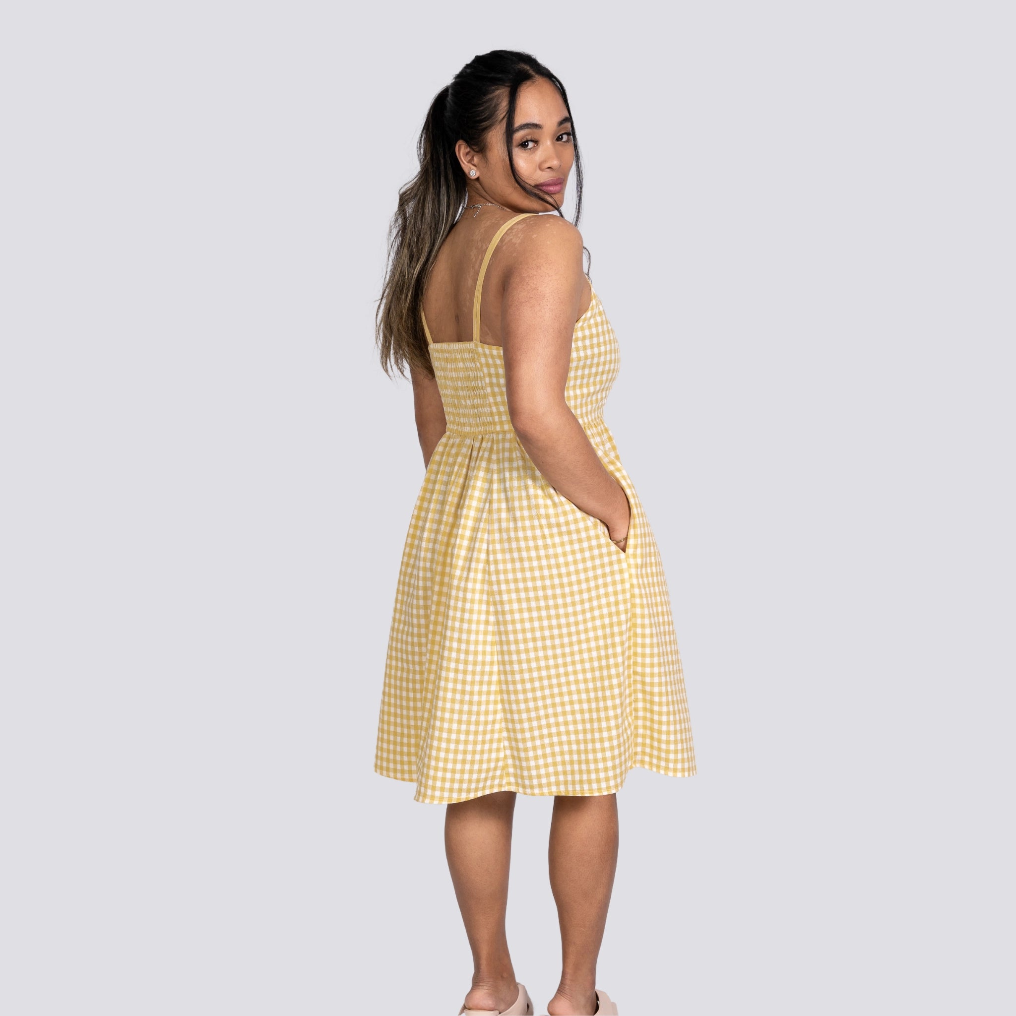 A woman in a Sunshine Chic Yellow Plaid Cotton Mini Dress by Karee looks over her shoulder, standing against a grey background.