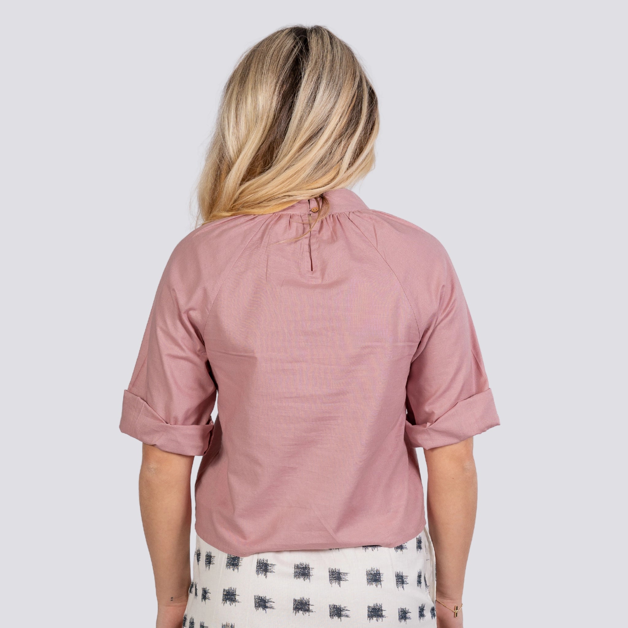 Rear view of a woman wearing a Karee Flamingo Rose 3/4 Sleeve Linen-Cotton Top with a buttoned keyhole closure. She has blonde hair and stands against a grey background.