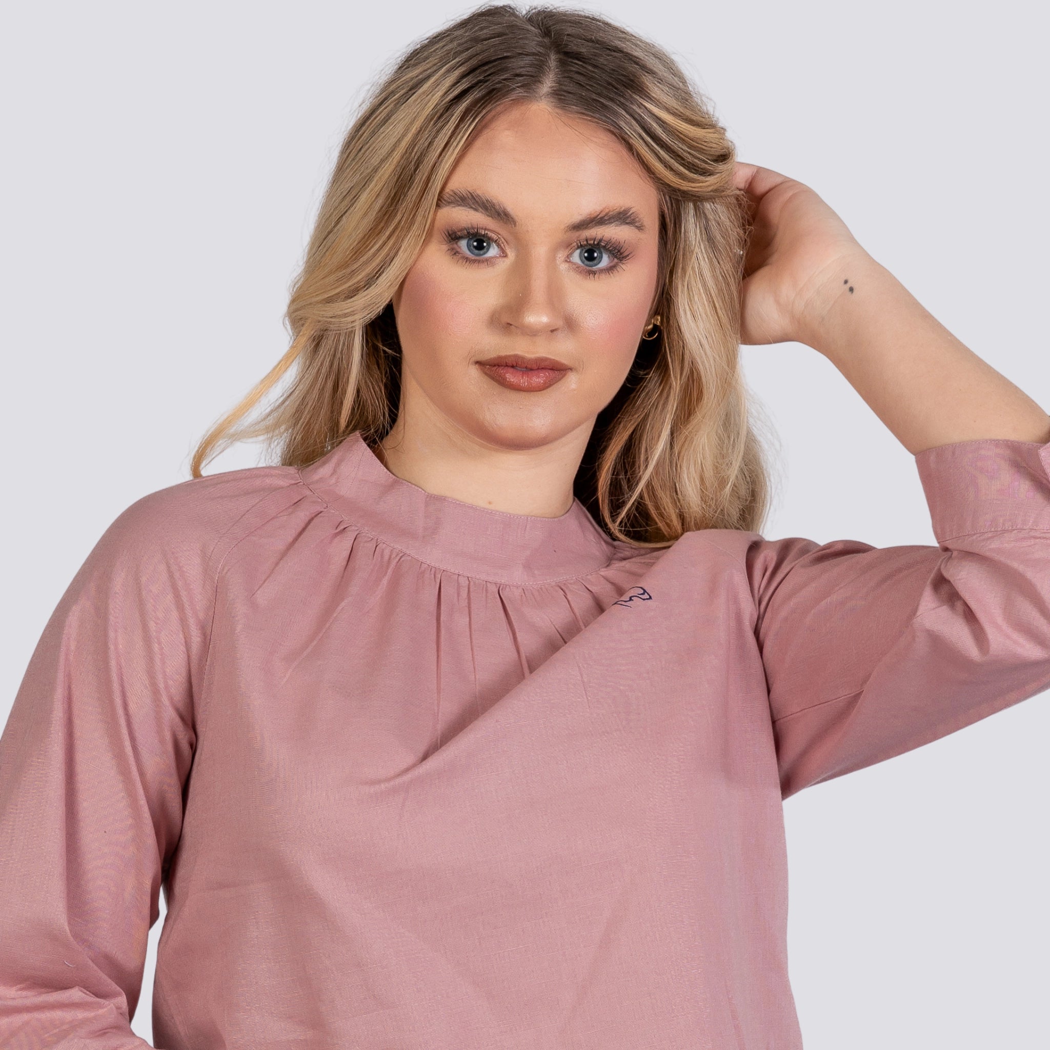 A woman with blonde hair poses, touching her head, wearing a Karee Flamingo Rose 3/4 Sleeve Linen-Cotton Top For Women with a high neckline.